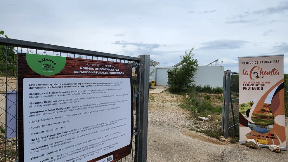Puerta de acceso metálica al Centro de Naturaleza La Chanta con un cartel informativo sobre las normas de conducta en espacios naturales protegidos y un tótem publicitario de Holcim y Brinzal.
