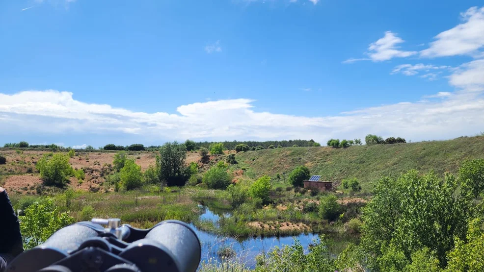 Prismáticos de observación fijos en primer plano enfocando el humedal protegido de La Chanta rodeado de vegetación mediterránea bajo un cielo azul con nubes.